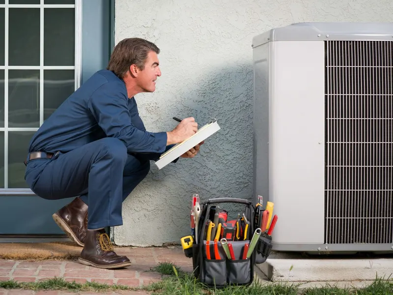 A GV's technician kneeling beside an outdoor air conditioning condenser, taking notes on a clipboard