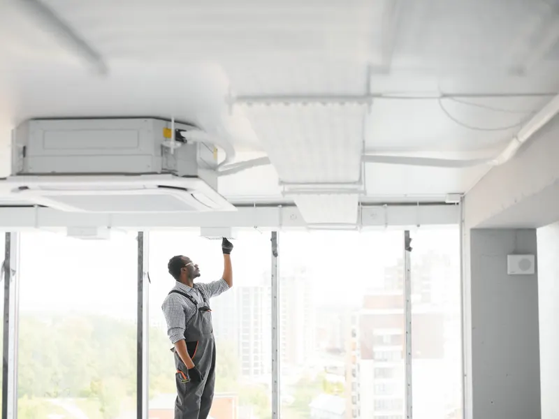 A technician on a ladder servicing a ceiling-cassette unit in a bright commercial space