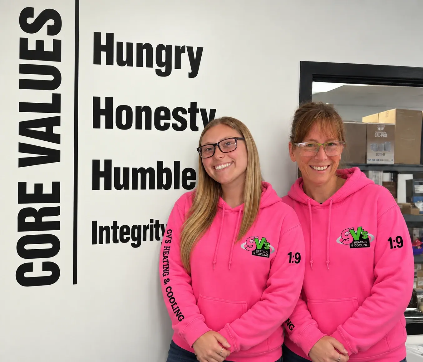 Two members of the GV's office team in pink hoodies beside the company core values wall.